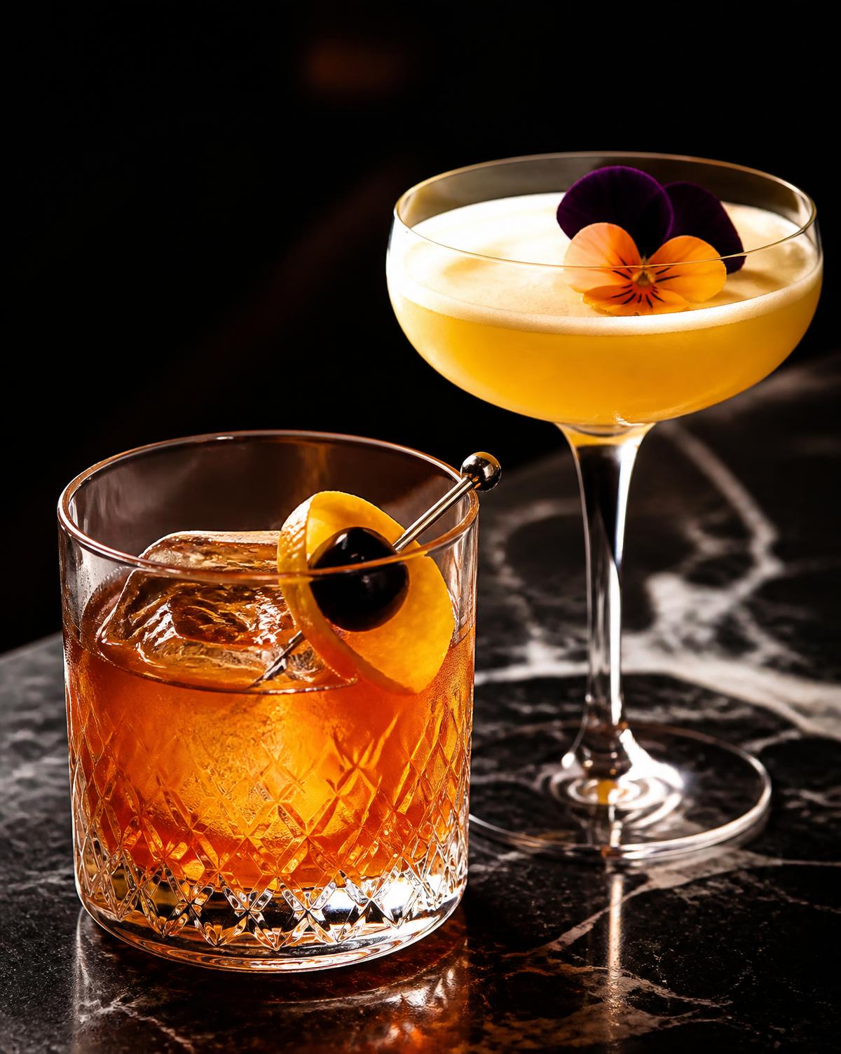 Two garnished craft cocktails on a dark marble bar
